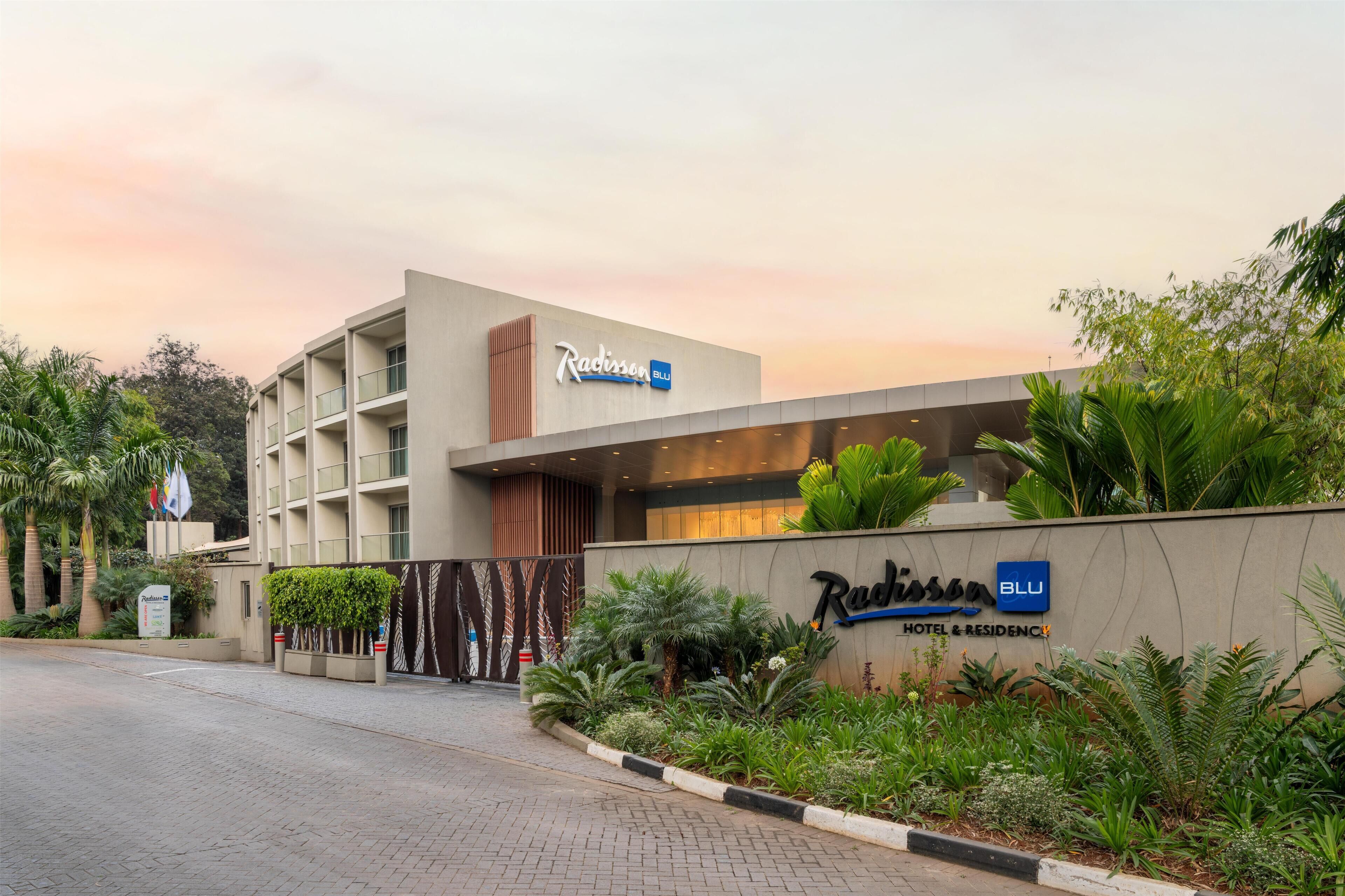A modern hotel entrance with a sign for Radisson Blu, surrounded by lush greenery and a paved driveway.