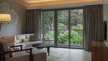 A modern hotel room with a sofa, armchair, and a small table. It features a large window with a view of greenery outside.