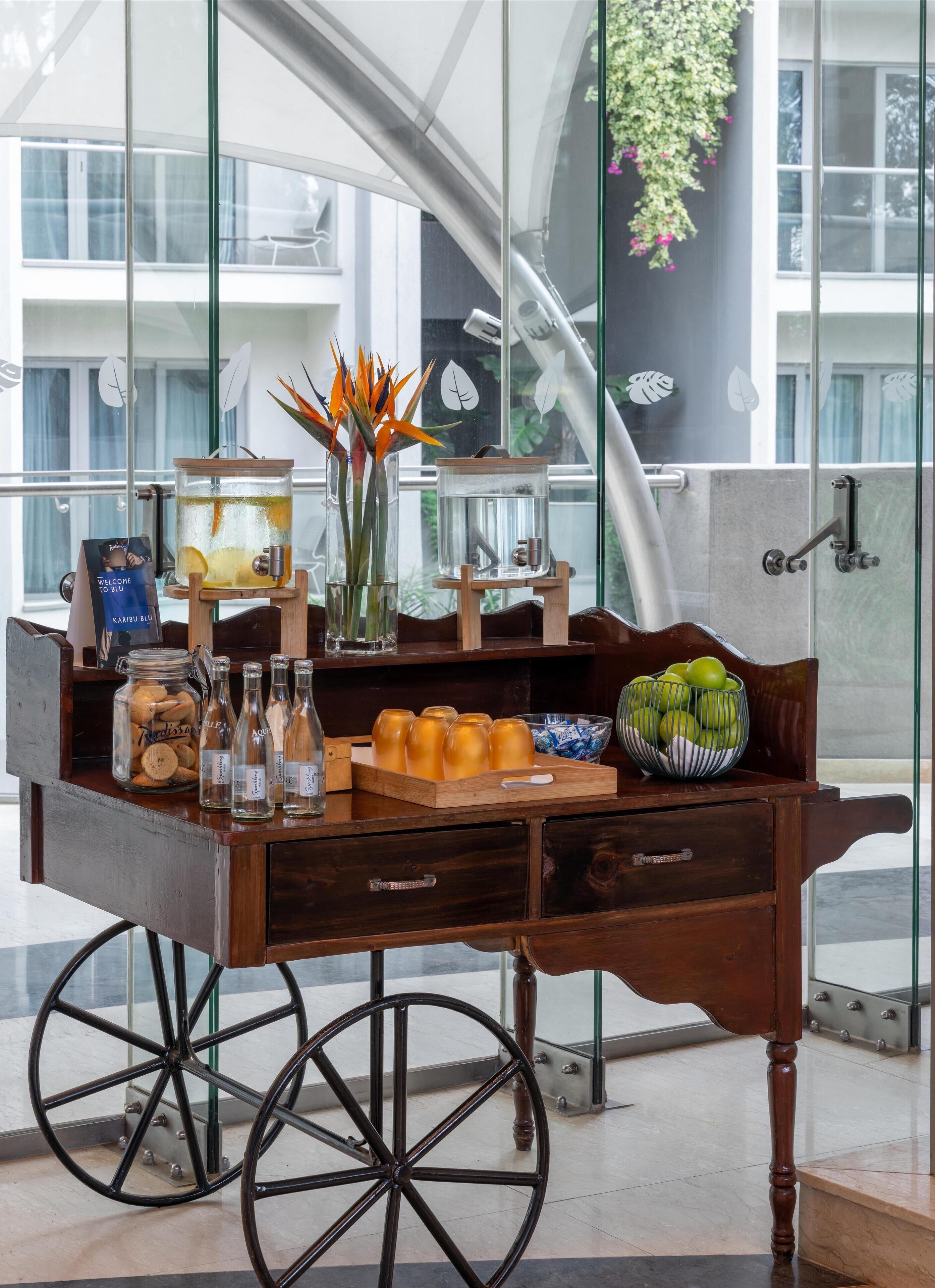 A wooden cart with a glass jar of almonds, several bottles, a bowl of apples, and a decorative plant.