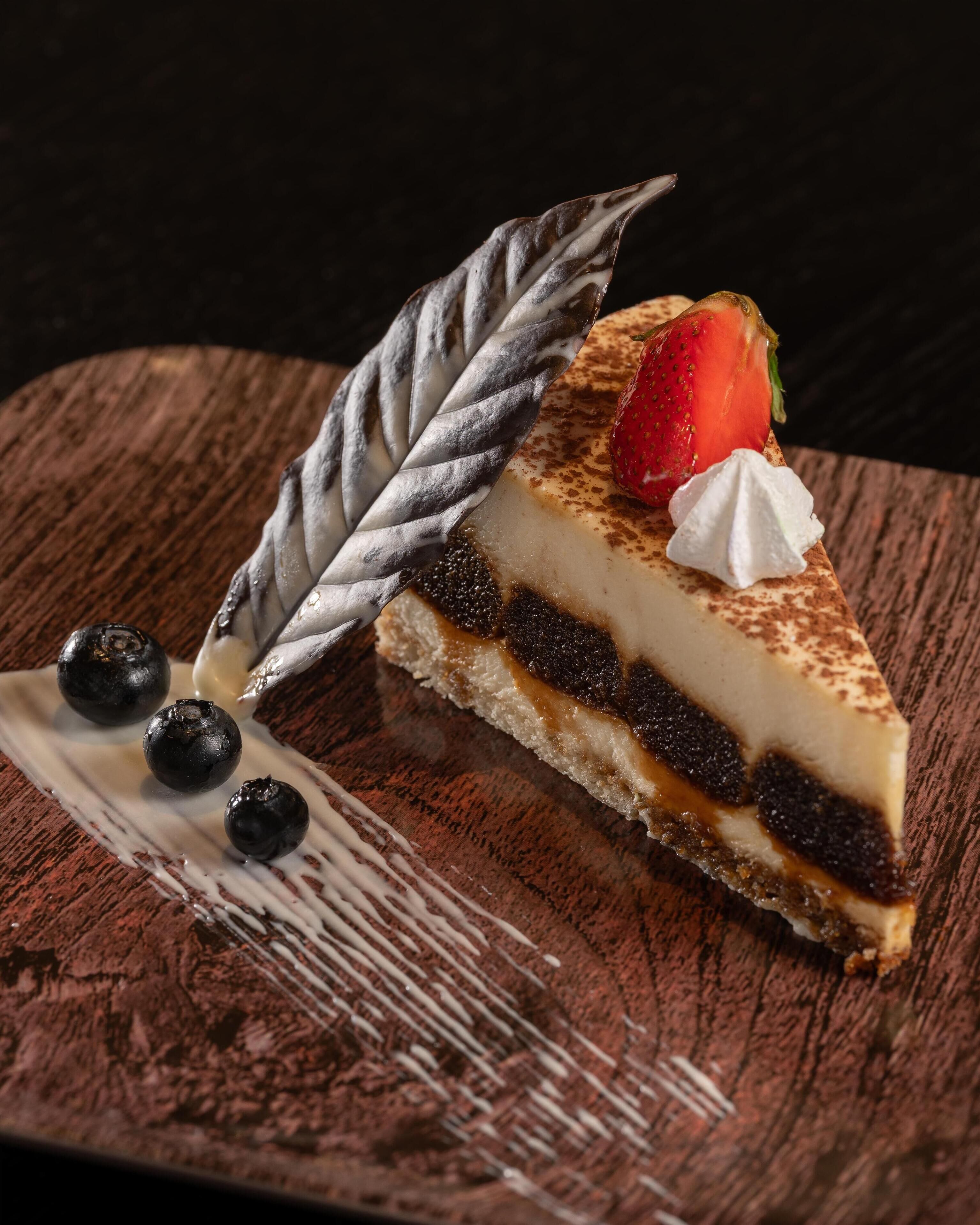 Une part de gâteau au fromage, garnie d’une fraise et d’une feuille décorative, présentée sur une planche en bois.
