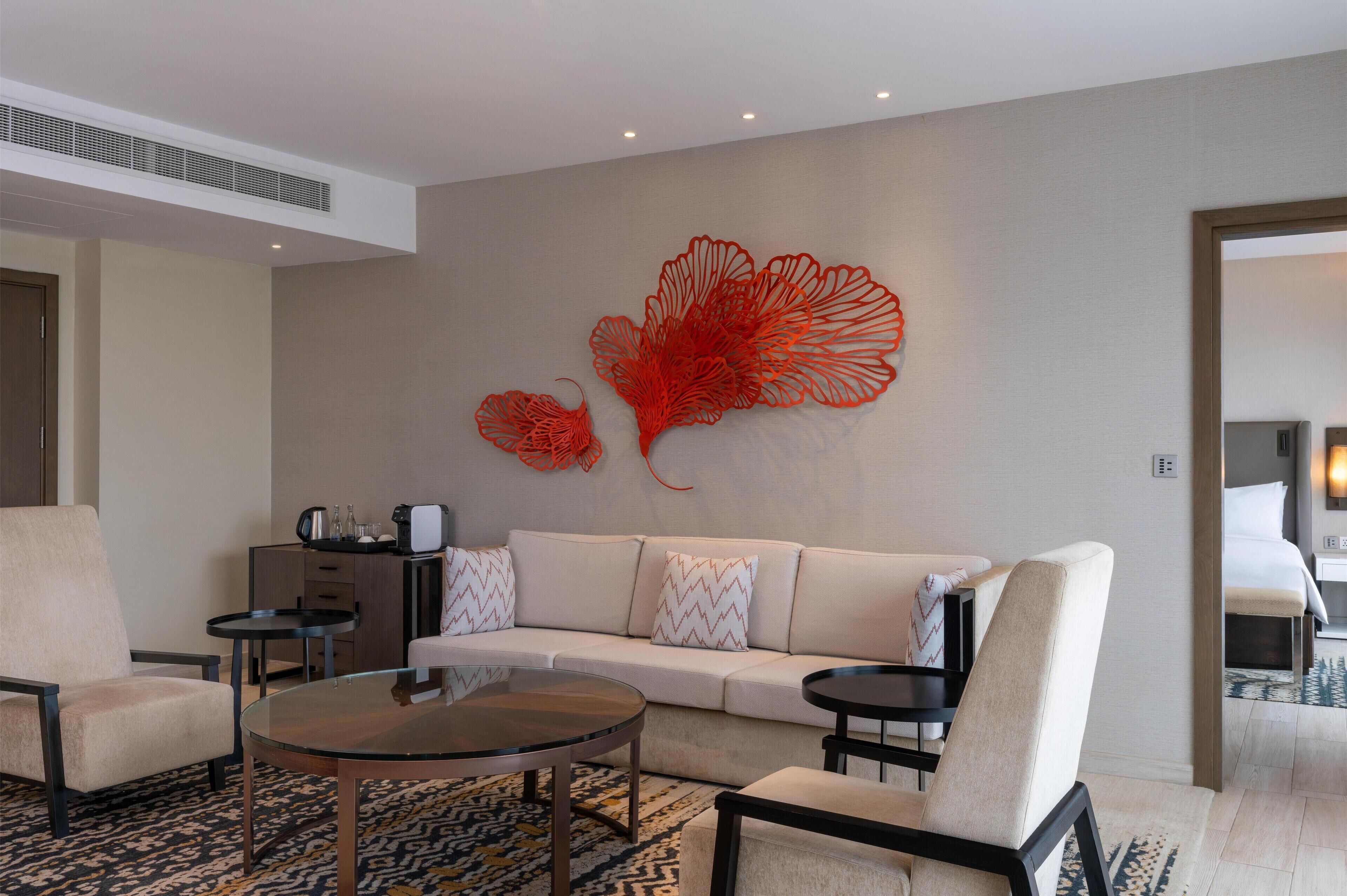 A modern hotel room with a beige sofa, two armchairs, a glass coffee table, and a decorative red wall piece.