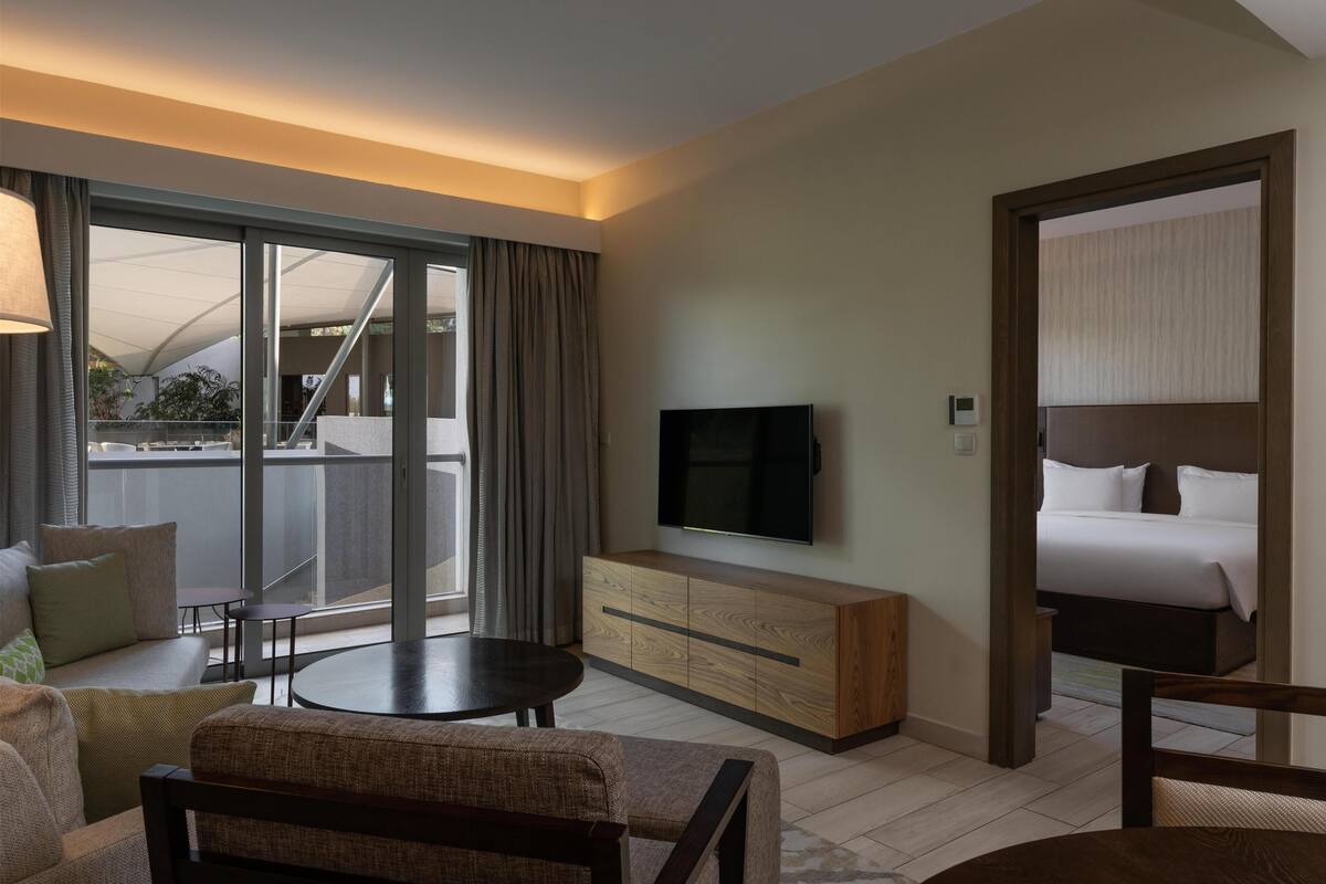 A hotel room with a large sliding glass door to a balcony, a flat-screen TV mounted on a wooden cabinet, a bed with white linens, and a small round table.
