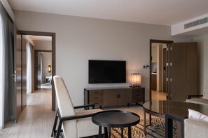 A modern hotel room with a flat-screen TV, a sofa, a glass coffee table, a wooden cabinet, and a patterned rug.