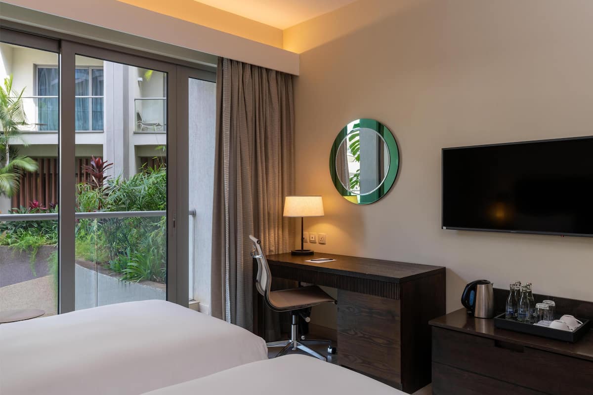 A hotel room with a bed, a desk with a chair, a TV, a lamp, and a view of a balcony with plants.