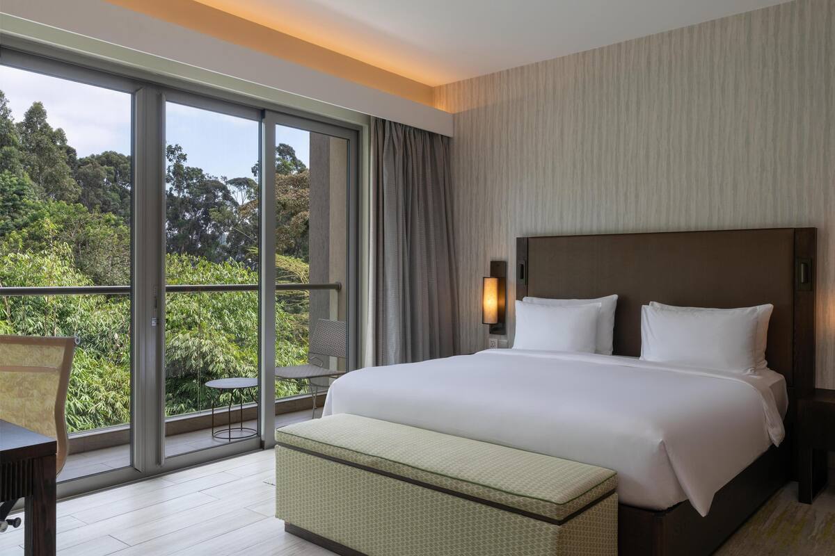 A hotel room with a large bed, a view of greenery through a sliding door, and a small table and chair by the window.