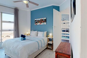 A bedroom with a bed, a ceiling fan, a window with curtains, a wall art piece, and a bunk bed in the adjacent room. - The Bay Views (Cape Town)