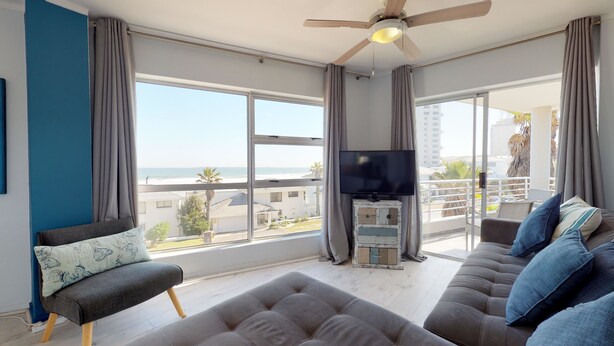 A modern living room with a large window offering a view of the beach and buildings, a ceiling fan, and a flat-screen TV on a wooden stand. - The Bay Views (Cape Town)