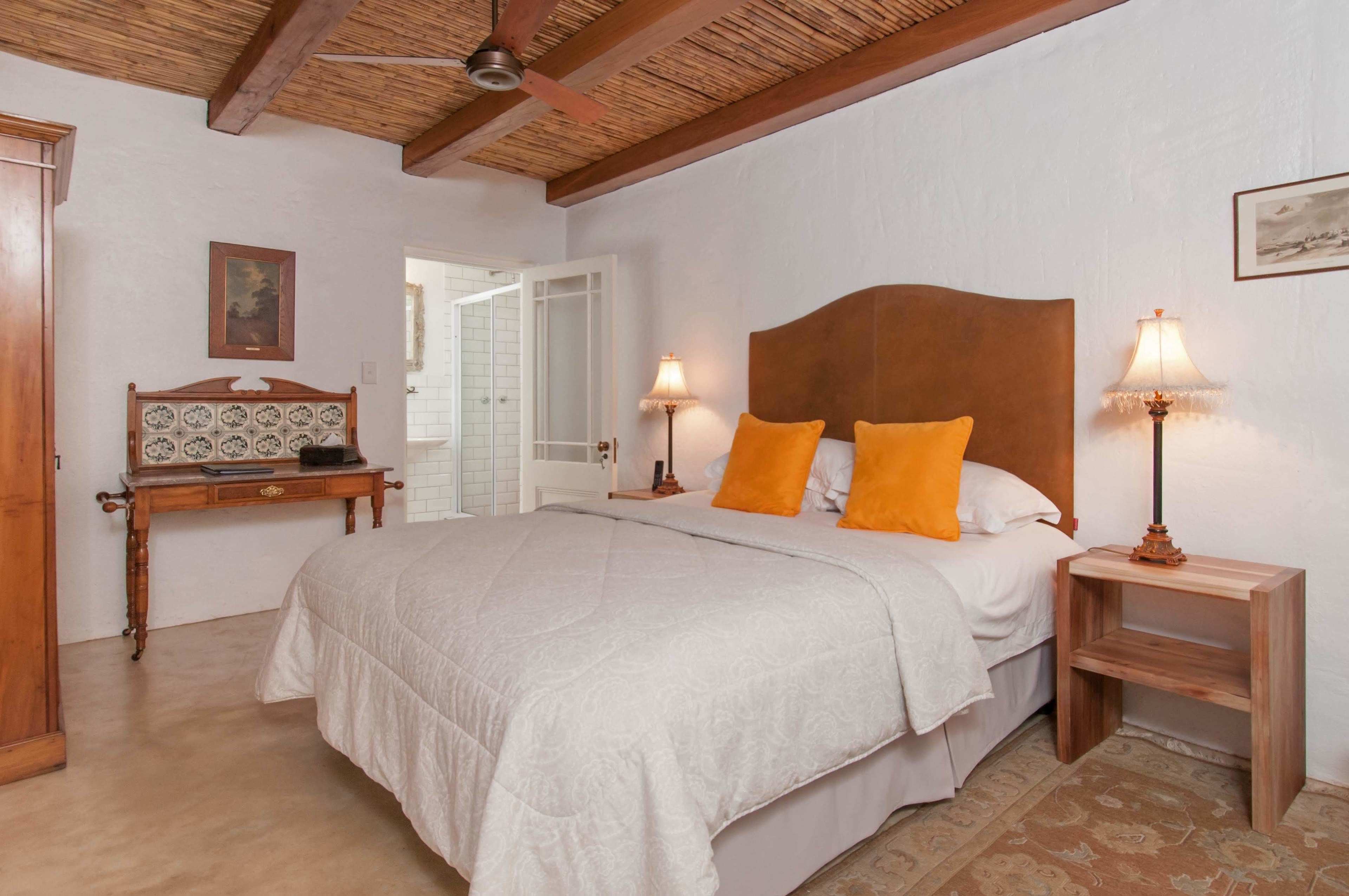 Une chambre à coucher avec un grand lit, deux tables de chevet avec des lampes, une tête de lit en bois, une petite table ornée d’un objet décoratif et une porte donnant sur une autre pièce.