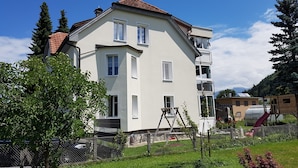Un immeuble résidentiel à plusieurs étages, doté d’un toit en pente, de balcons et d’une cour clôturée avec des arbres et une aire de jeux.