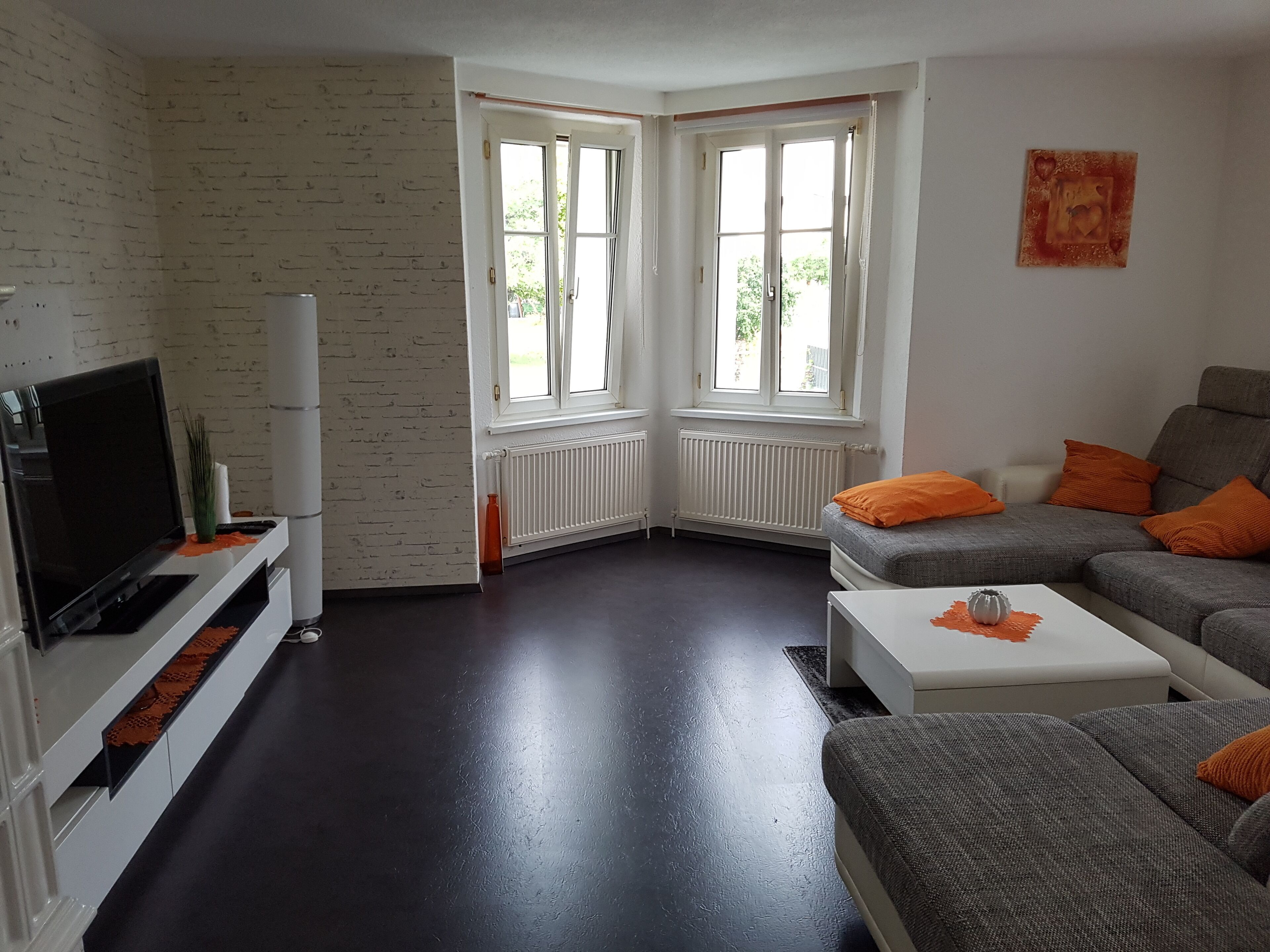 A modern living room with a flat-screen TV, grey sofa, orange cushions, and a white coffee table.