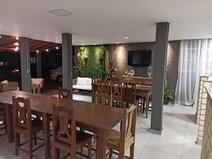 A dining area with a long wooden table and chairs, a sofa, a flat-screen TV mounted on the wall, and a green wall with plants. - Residencial Encantos do Rosa (Imbituba)