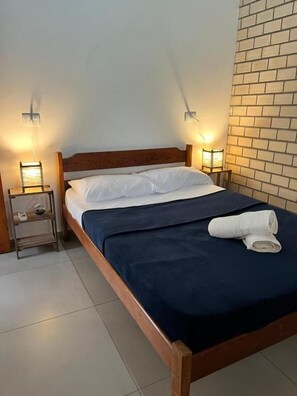 A single bed with a wooden headboard, a dark blue bedspread, and a white pillow. There are two bedside tables with lamps, and a brick wall to the right. - Residencial Encantos do Rosa (Imbituba)