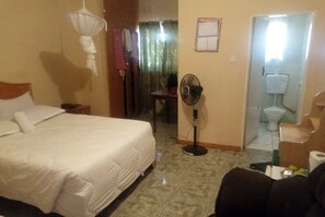 A hotel room with a large bed, a standing fan, a small table, and a bathroom visible through an open door.