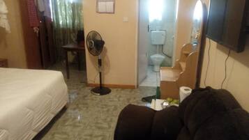 A hotel room with a bed, a dark sofa, a standing fan, a TV, and a bathroom visible through an open door.