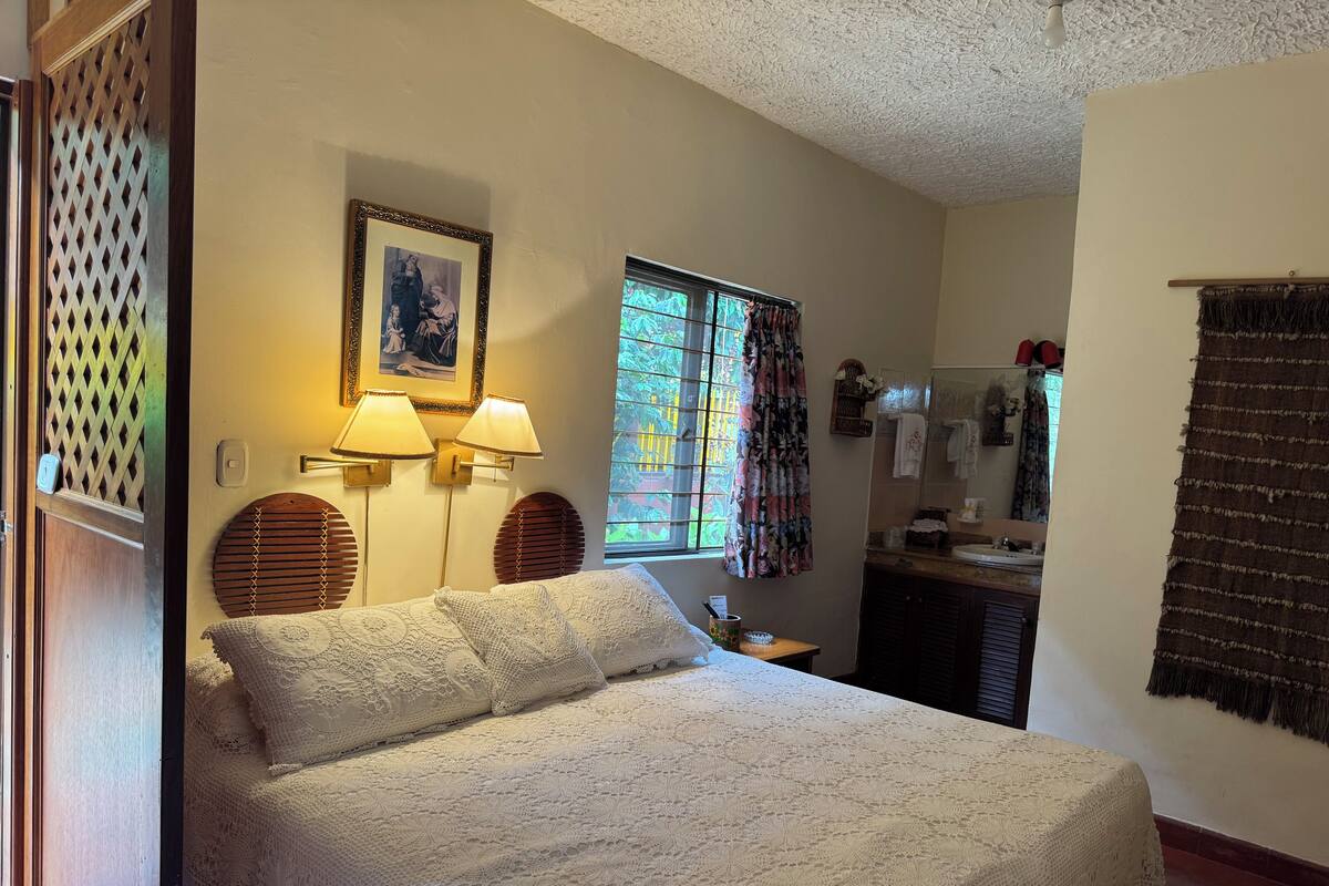 A bedroom with a large bed, two wall-mounted lamps, a window with curtains, a mirror, and a small table with a lamp.