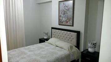 A bedroom with a bed, a nightstand, a bedside table, a framed picture on the wall, and a window with curtains.