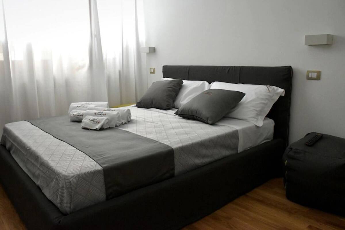 Una cama bien tendida con un edredón gris y blanco, dos almohadas y un control remoto sobre una mesita de noche.