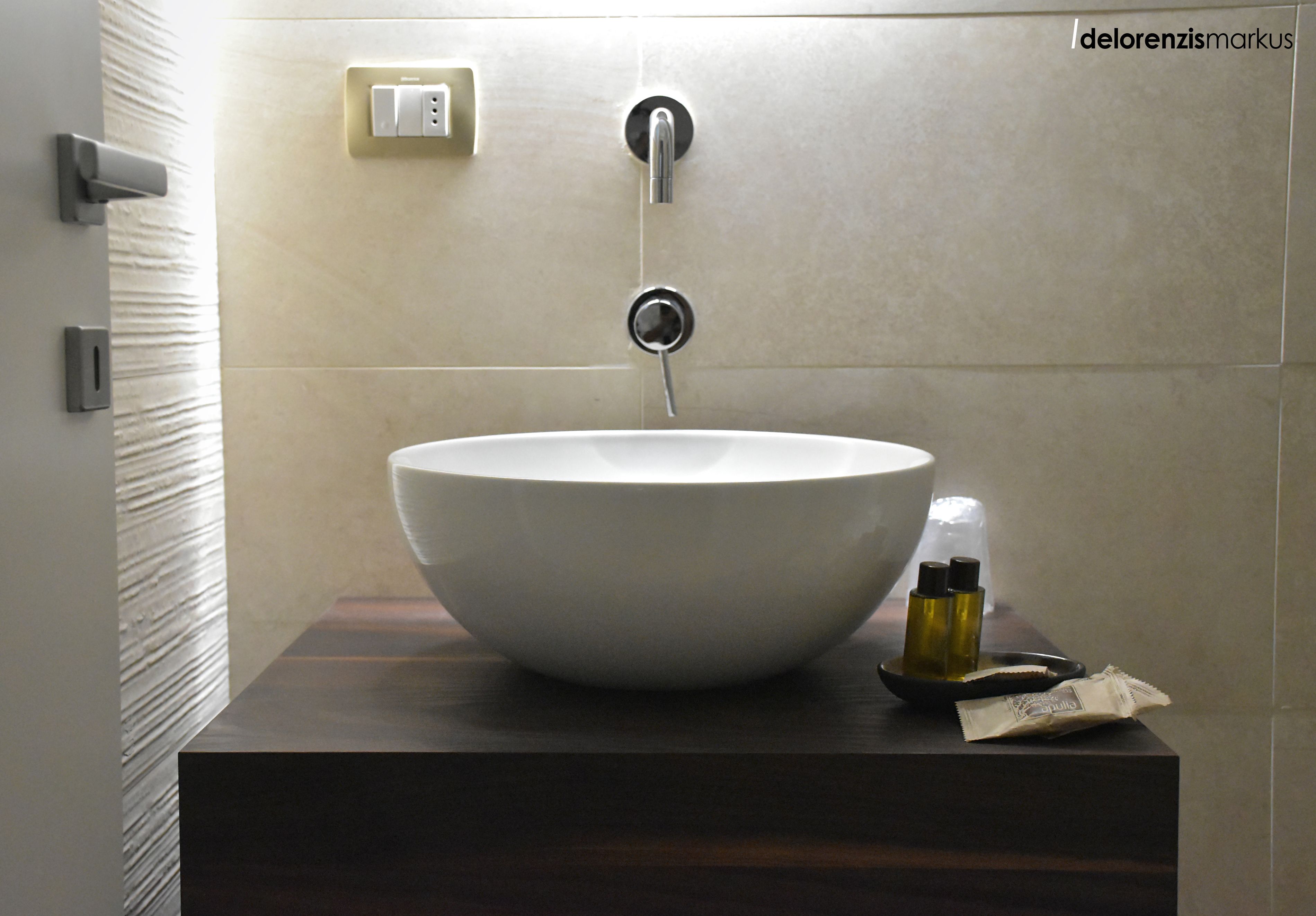 Un baño con un lavamanos blanco de diseño, un mueble de tocador de madera oscura, un dispensador de toallas y una botella de jabón líquido.