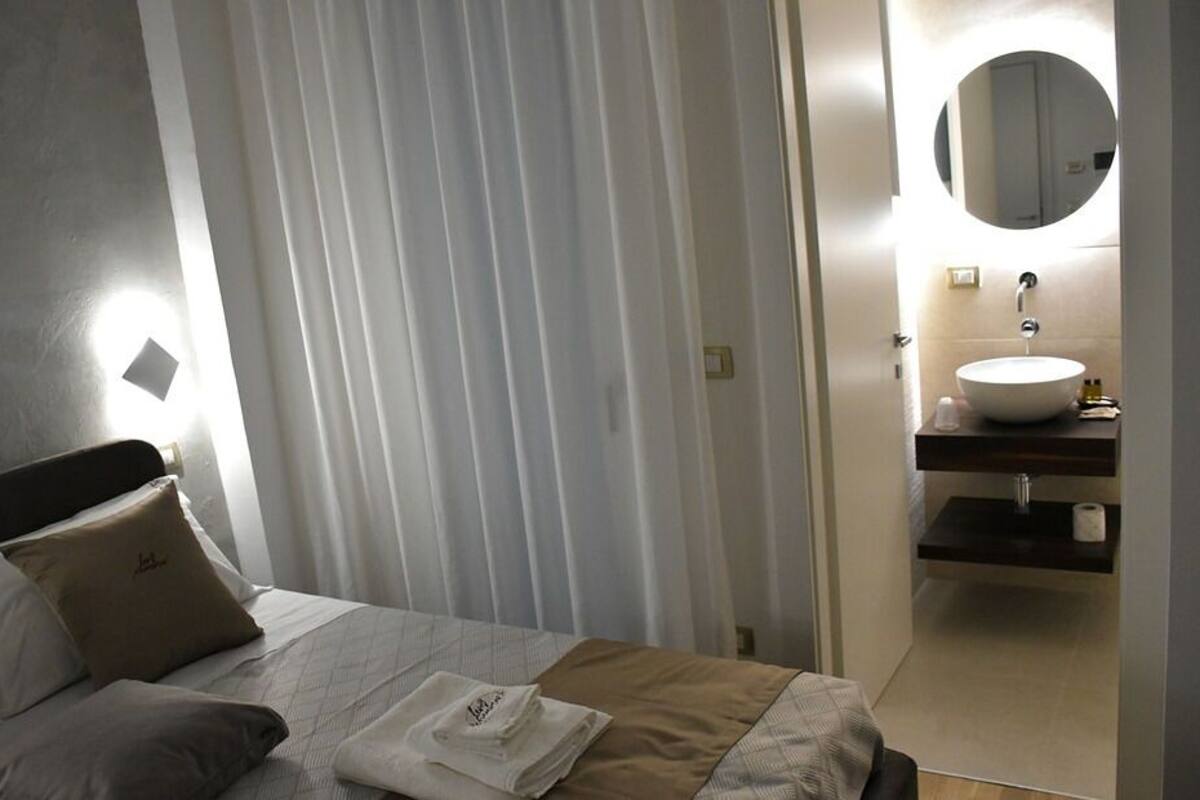 Habitación de hotel con cama, mesita de noche, espejo redondo y baño con lavamanos e inodoro.