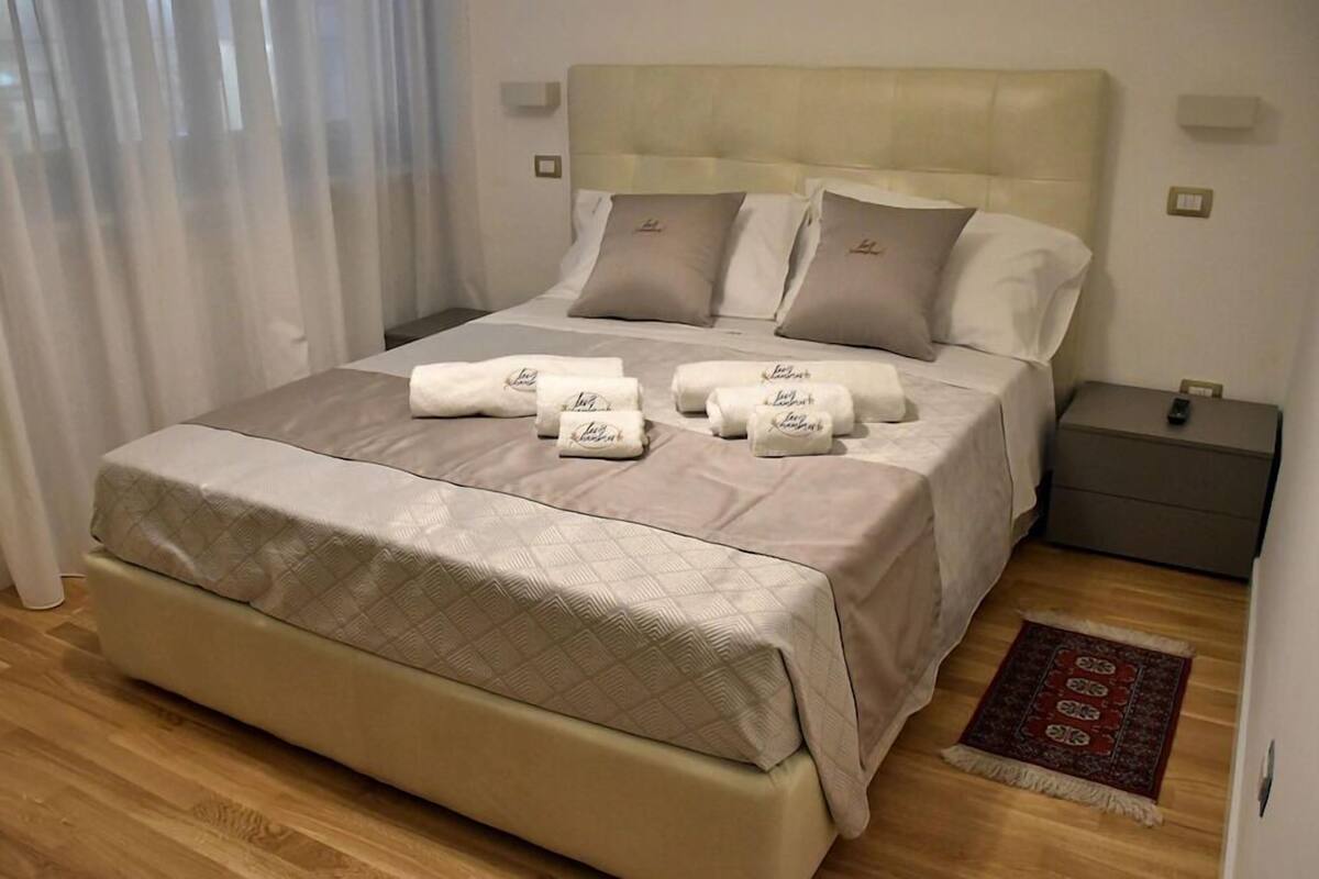 Una cama bien hecha con un cabecero color crema, una mesita de noche con una lámpara y una alfombra pequeña en el suelo.