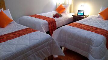 Habitación de hotel con tres camas individuales, cada una con ropa blanca y detalles en naranja. Hay un ordenador portátil sobre una mesita de noche, y una pequeña mesa con una lámpara y una flor.