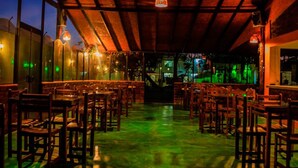 An indoor dining area with wooden tables and chairs, a high ceiling with exposed beams, and ambient lighting.