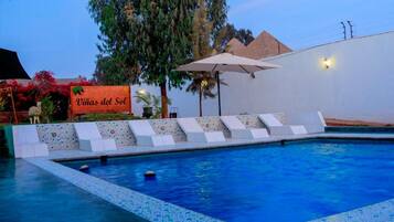 Una piscina con borde de azulejos, un área de descanso con tumbonas blancas y un letrero que dice "Villas del Sol".