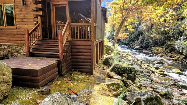 A wooden cabin with a porch and steps leading to a door, surrounded by a rocky stream and autumn foliage. - Whitewater Mountain Cabin on the Cullasaja River  (Highlands)