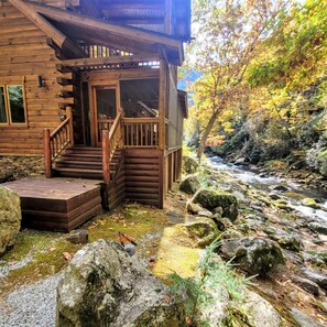 Una cabaña de madera con un porche y escalones que conducen a una puerta, rodeada por un arroyo pedregoso y follaje otoñal.