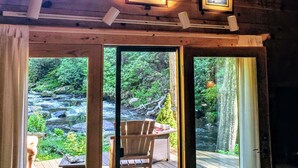 A wooden interior with framed artwork, a view of a natural landscape through large windows, and a deck area with outdoor seating. - Whitewater Mountain Cabin on the Cullasaja River  (Highlands)