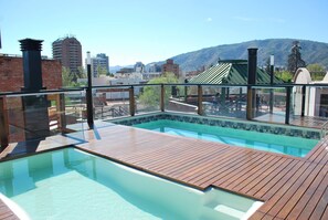 An outdoor swimming pool with wooden decking, surrounded by a glass railing, overlooking a cityscape and mountains.