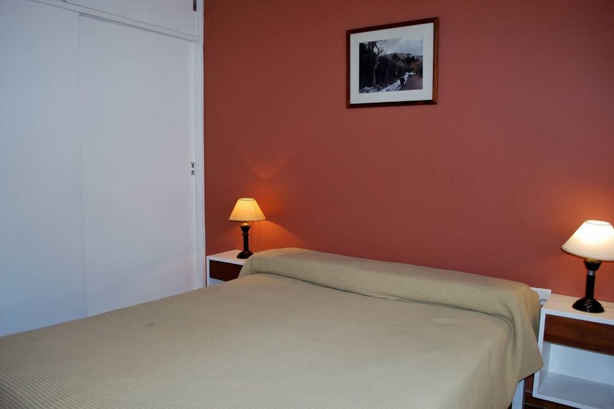 Une chambre à coucher avec un lit, deux tables de chevet avec des lampes, un tableau encadré au mur et une armoire blanche.