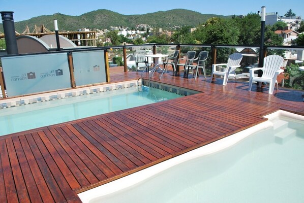 A wooden deck with a pool, white chairs, and a view of the city and hills in the background.