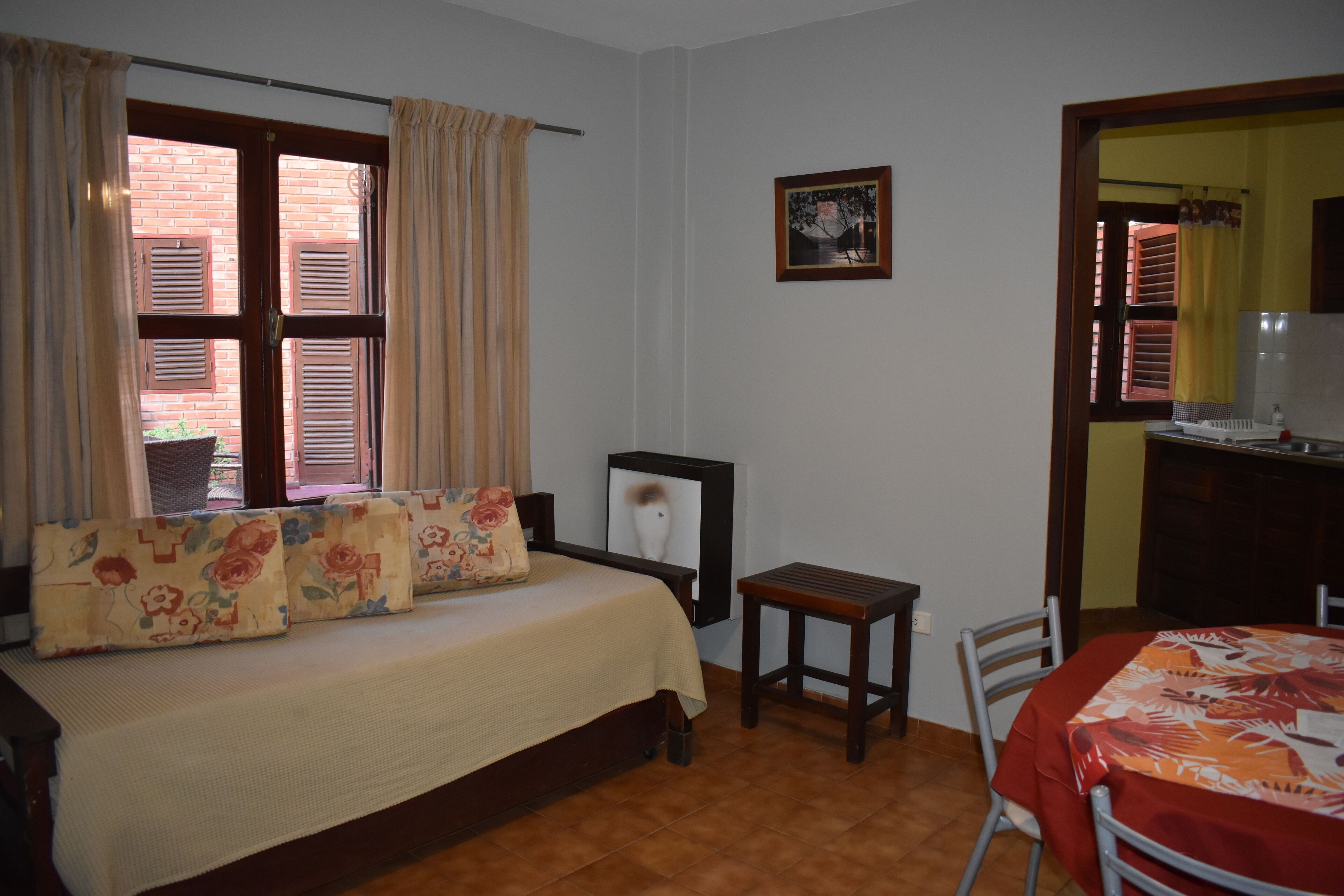 Una habitación con una cama, una mesita y una silla. Hay una ventana con contraventanas y un marco de fotos en la pared.