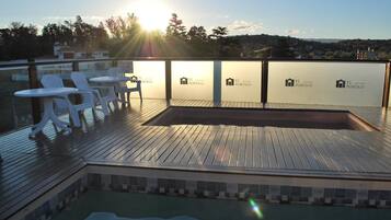 A rooftop pool area with white plastic chairs and tables, a wooden deck, and a view of the cityscape.