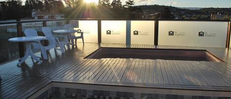 A rooftop pool area with white plastic chairs and tables, a wooden deck, and a view of the cityscape.