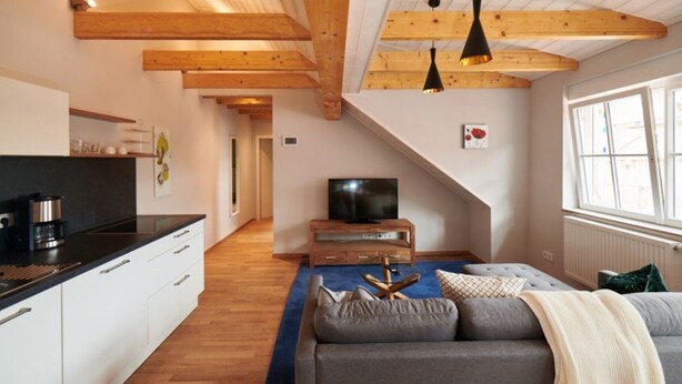 A modern loft-style room with exposed wooden beams, a kitchenette, a sofa, and a television.