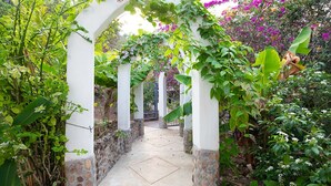 Un sentier de jardin bordé d’arches blanches et de socles en pierre, entouré d’une végétation luxuriante et de plantes en fleurs.