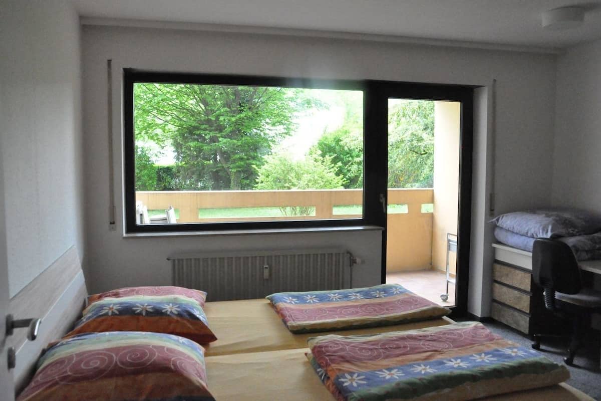 Ein Schlafsaalzimmer mit zwei Betten, einem Schreibtisch und einem Fenster mit Blick auf einen Balkon mit Bäumen.