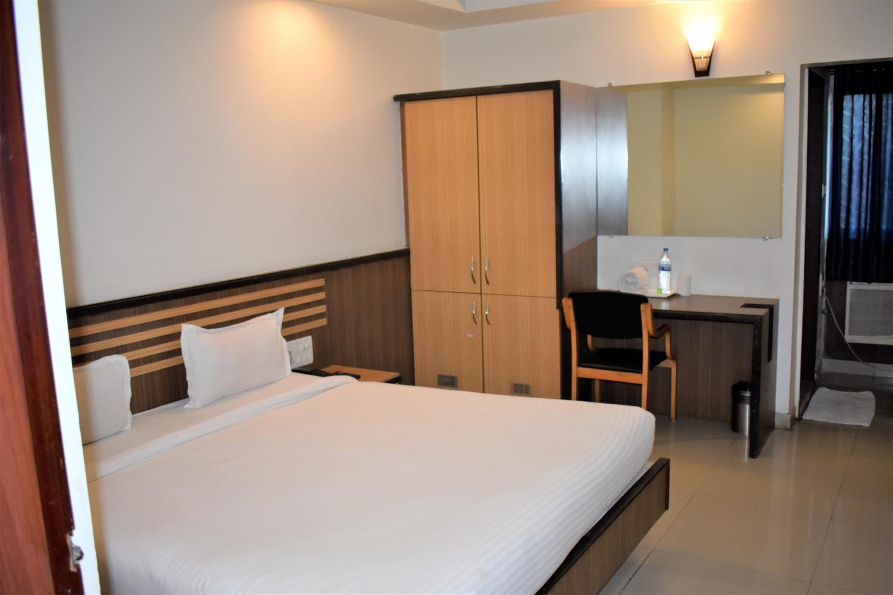 A hotel room with a large bed, a wooden wardrobe, a desk with a chair, a mirror, and a window with curtains.