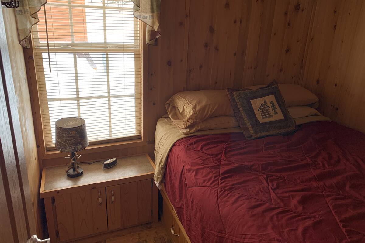 A wooden room with a bed, a window with blinds, a lamp, and a small table.