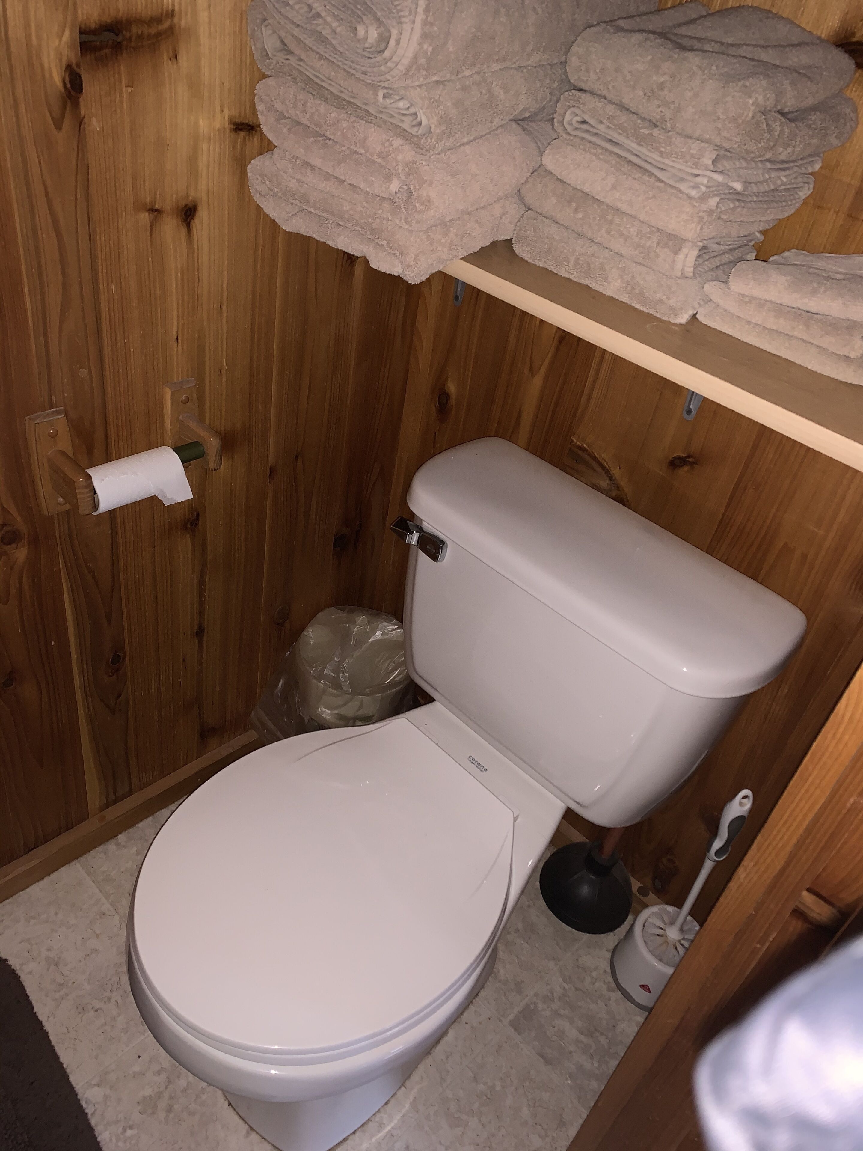 A wooden bathroom with a toilet, a roll of toilet paper, a plunger, and a stack of towels on a shelf.