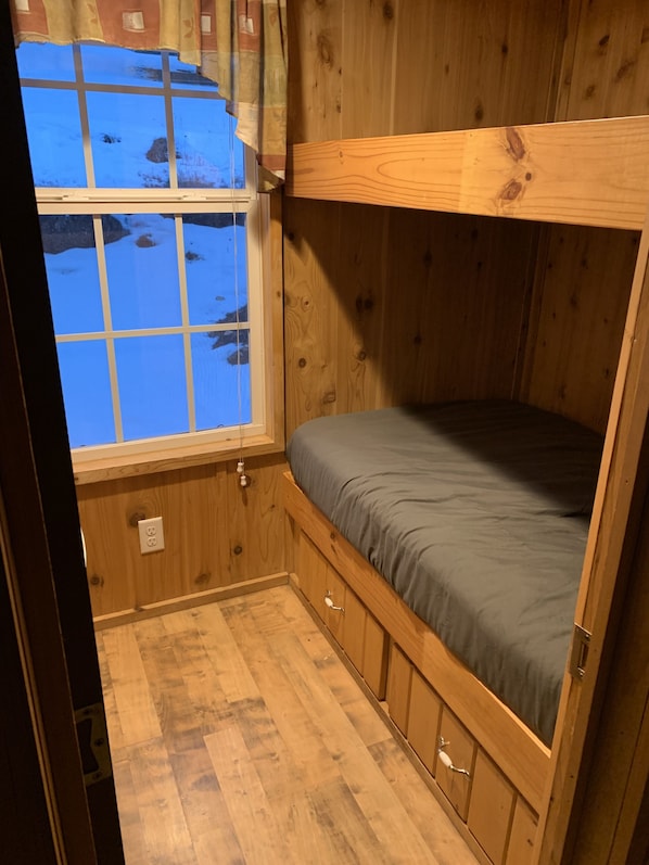 Una pequeña habitación tipo cabaña de madera con una cama nido, una ventana con cortinas y suelo de madera.
