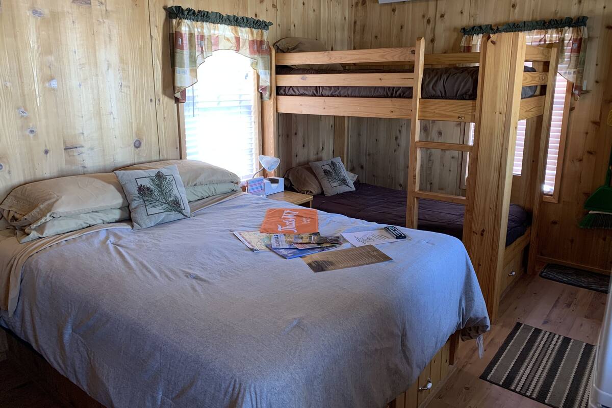 A wooden cabin room with a bunk bed, a desk with a lamp, and a window with curtains.