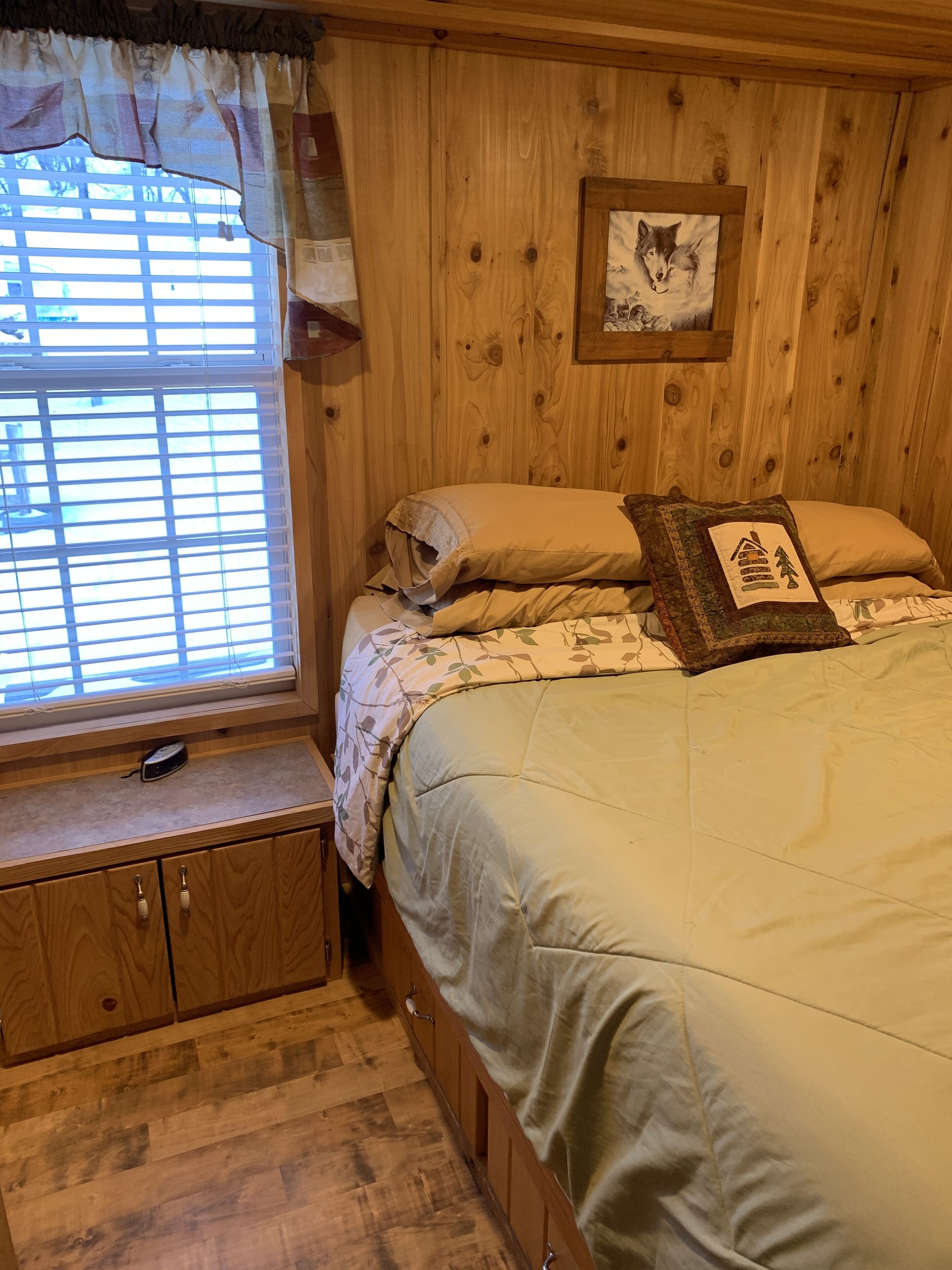 A wooden cabin-style bedroom with a large bed, a window with blinds, a small wooden cabinet, and a framed picture on the wall.