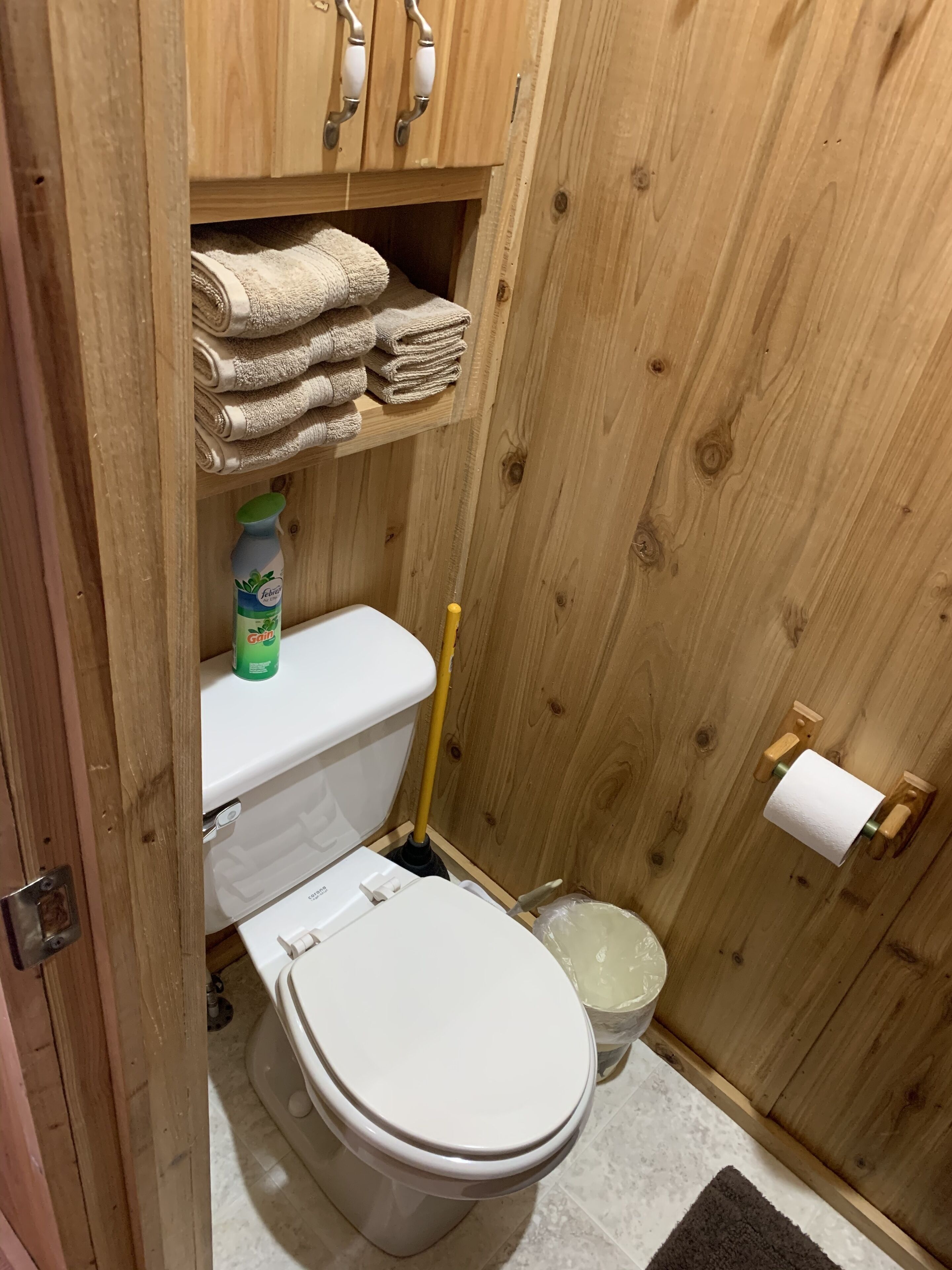 Un baño de madera con inodoro, un estante con toallas dobladas, una botella de producto de limpieza, un trapeador y un dispensador de papel higiénico.