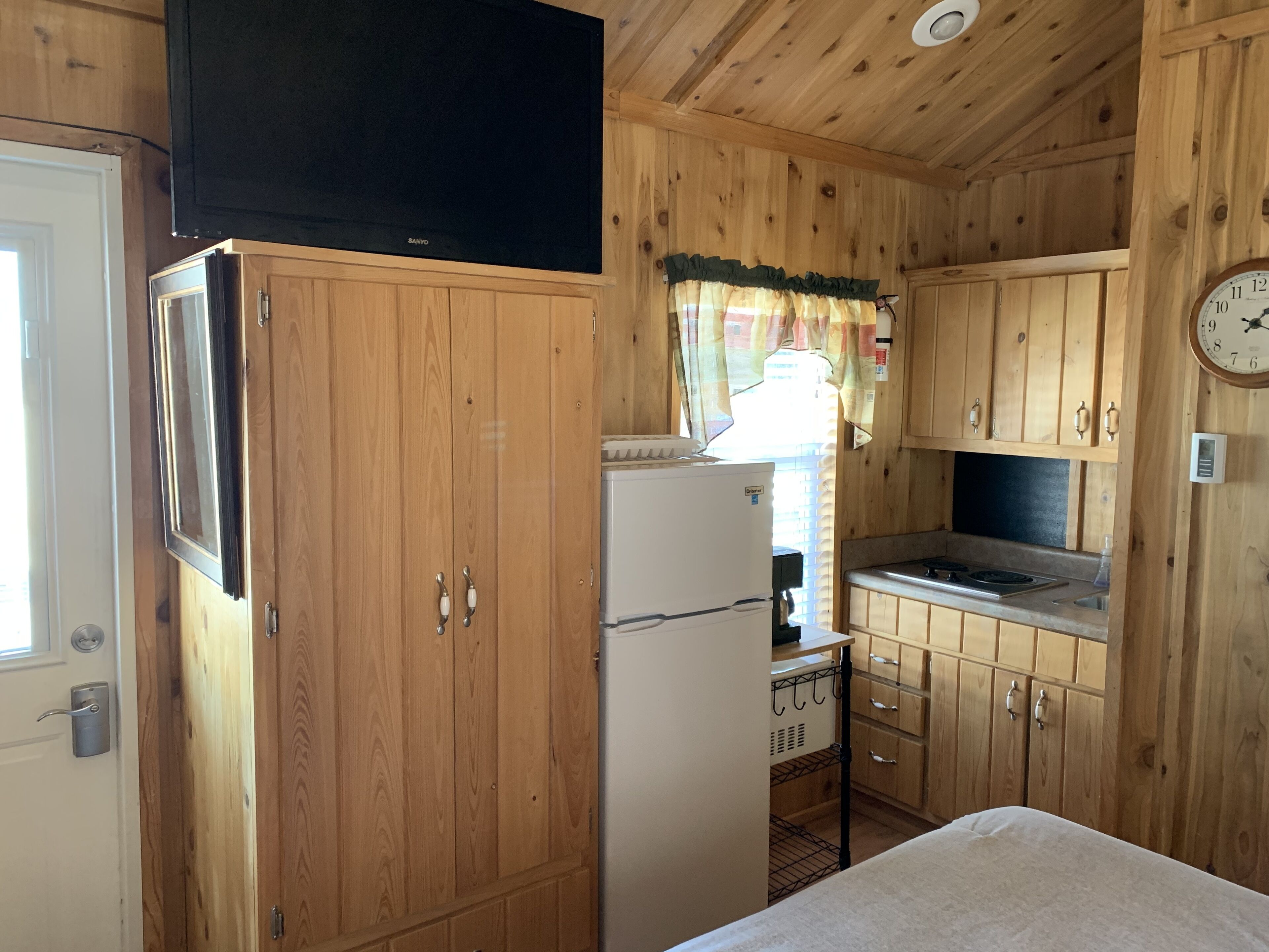 A compact kitchen with wooden cabinets, a refrigerator, and a clock on the wall.