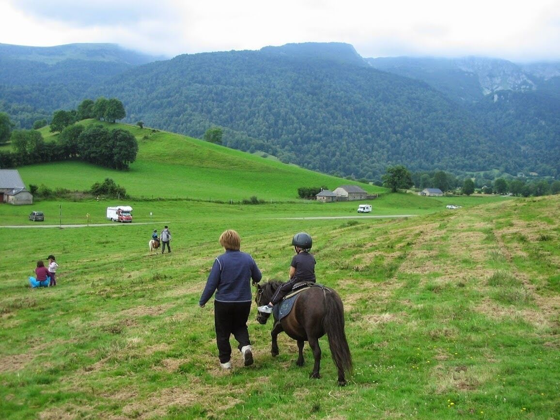 A rural landscape with a person riding a horse, another person walking beside, and a few others in the background.