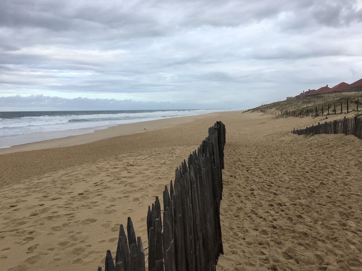 Ein sandiger Strand mit einem Holzzaun im Vordergrund und einem bewölkten Himmel.