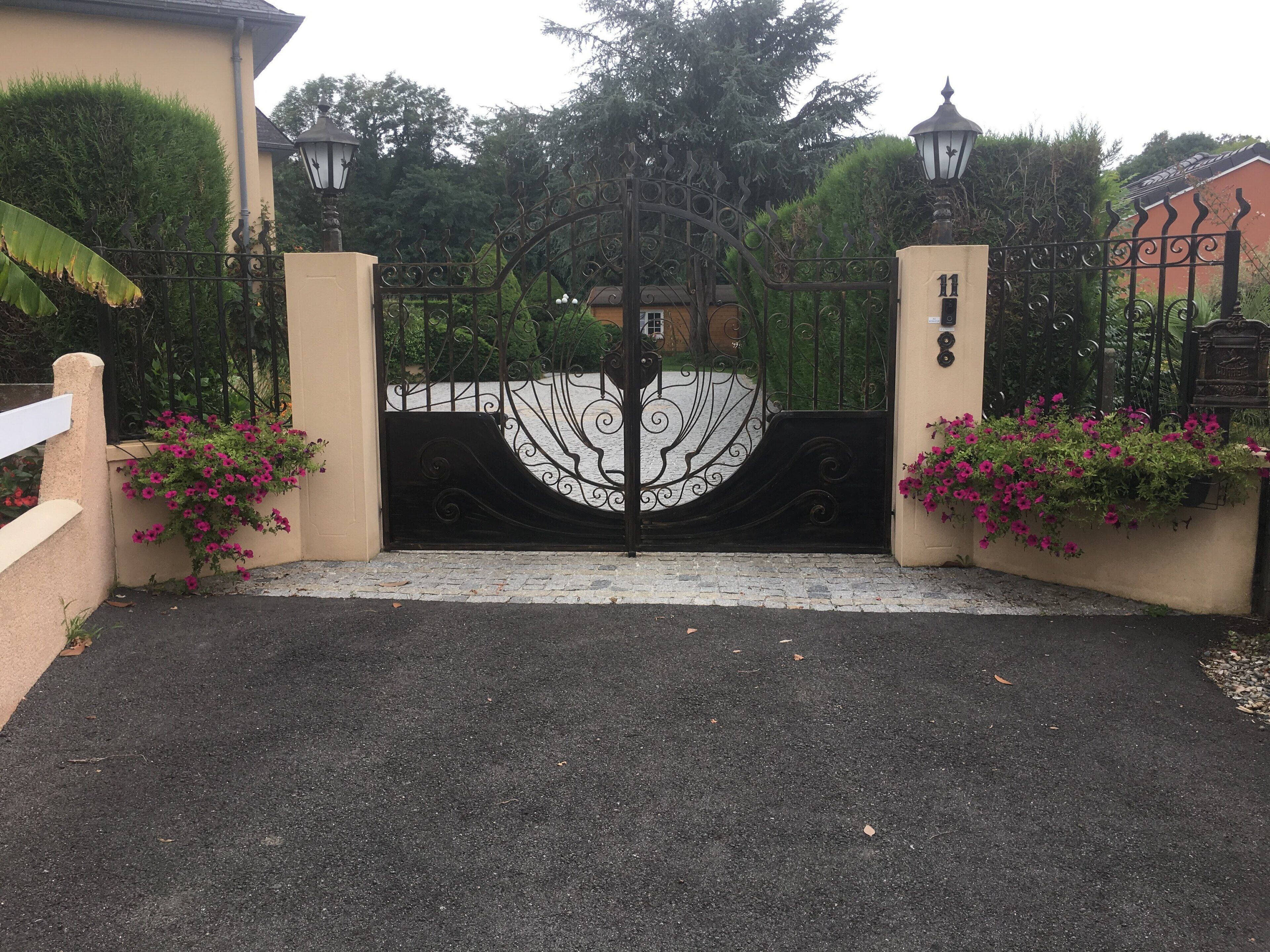 A gated entrance with a decorative iron gate, a paved driveway, and flower boxes with pink flowers.